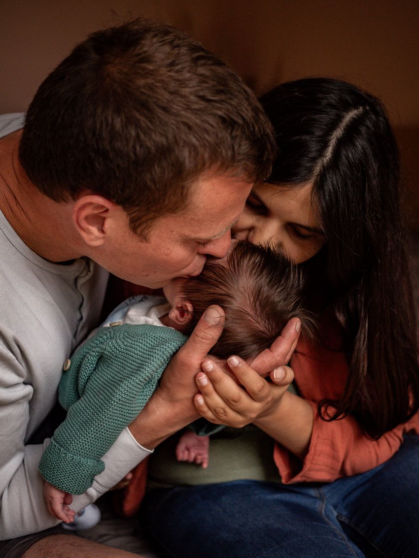 Newborn Shooting Ein Elternpaar hält sein Neugeborenes mit den Händen vor sich und küsst es liebevoll auf den Kopf