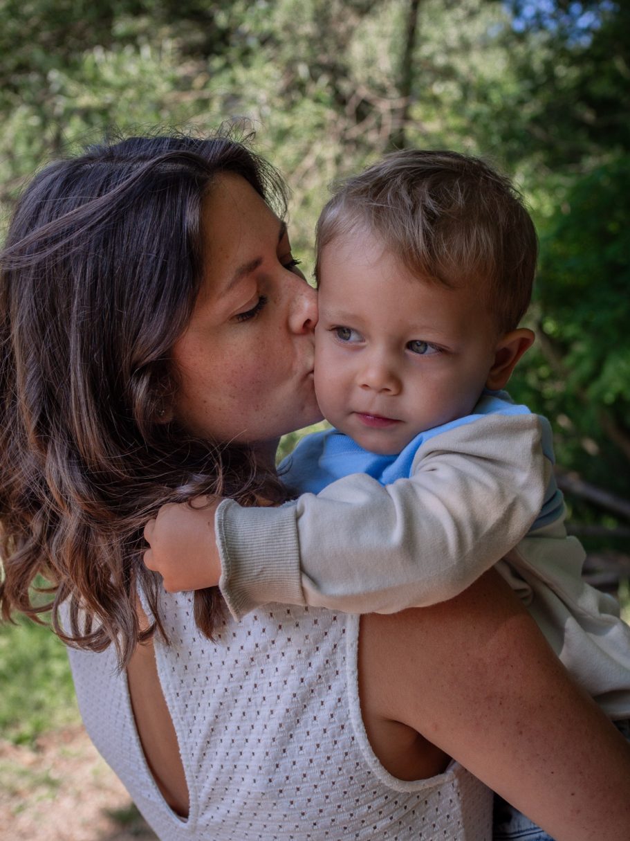Familienfotoshooting Eine Mutter küsst ihr kleines Kind, beide lächeln in einer natürlichen Umgebung.